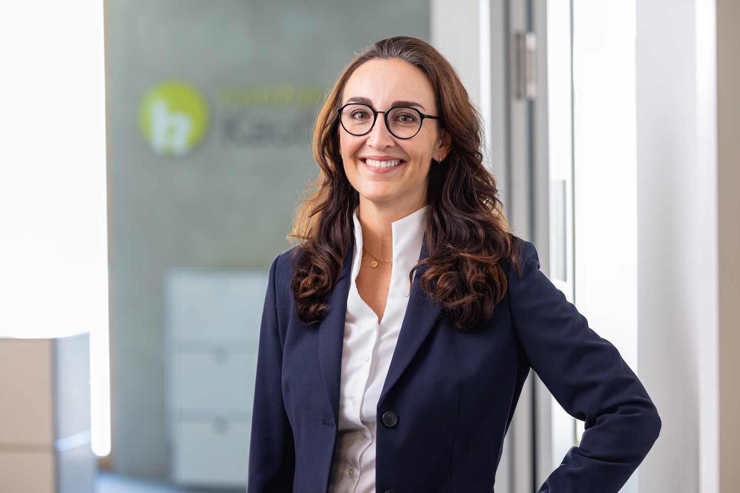 Porträt einer Frau im Blazer im Büro vor Glaswand mit Logo.