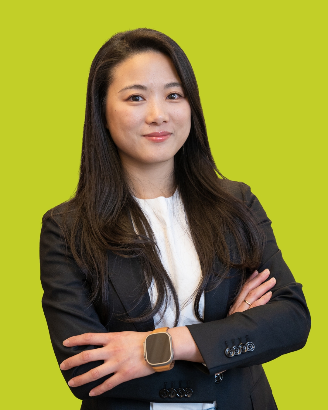 Porträt einer Frau im dunklen Blazer mit verschränkten Armen vor grünem Hintergrund.