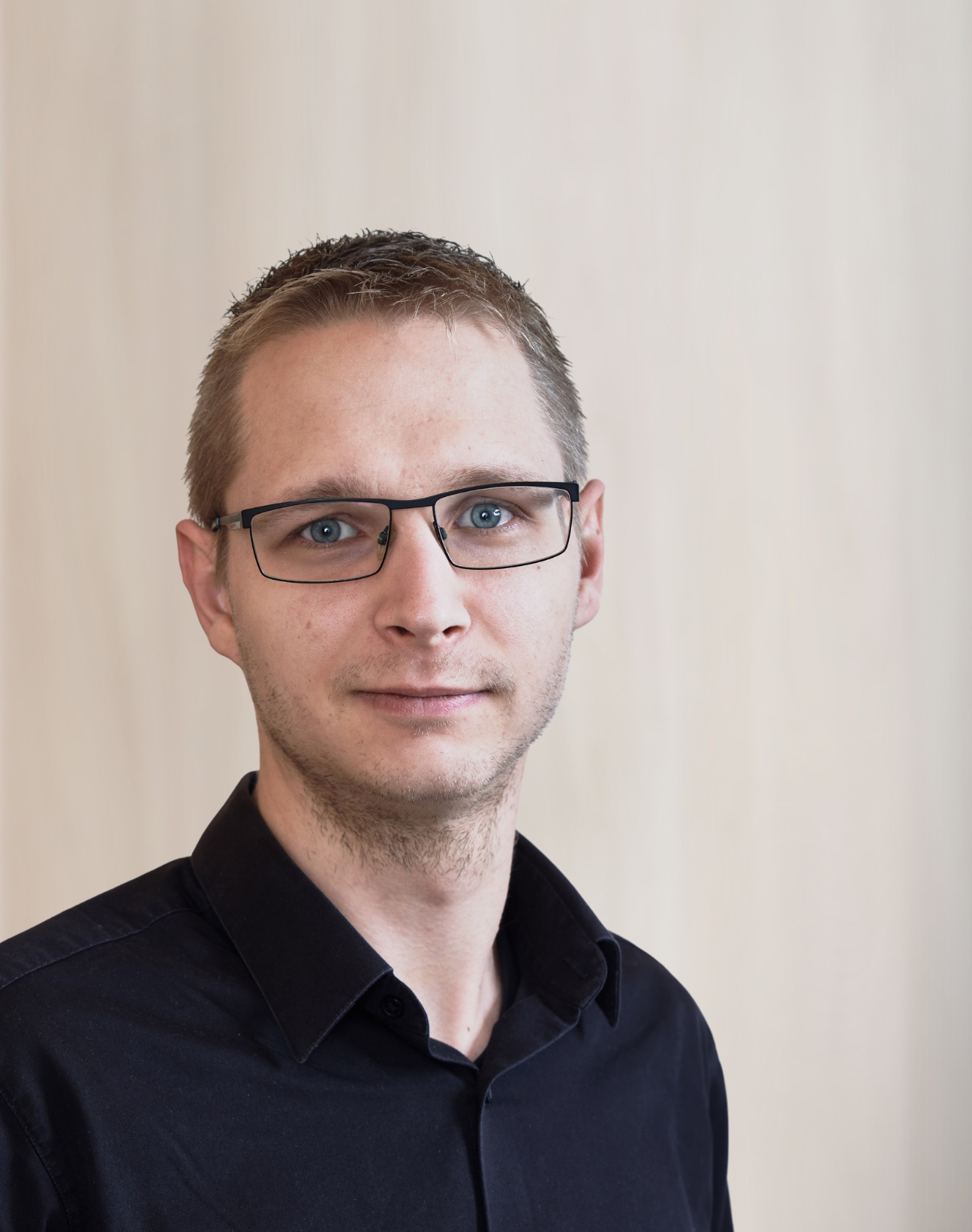 Porträt eines Mannes mit Brille im dunklen Hemd vor hellem Hintergrund.