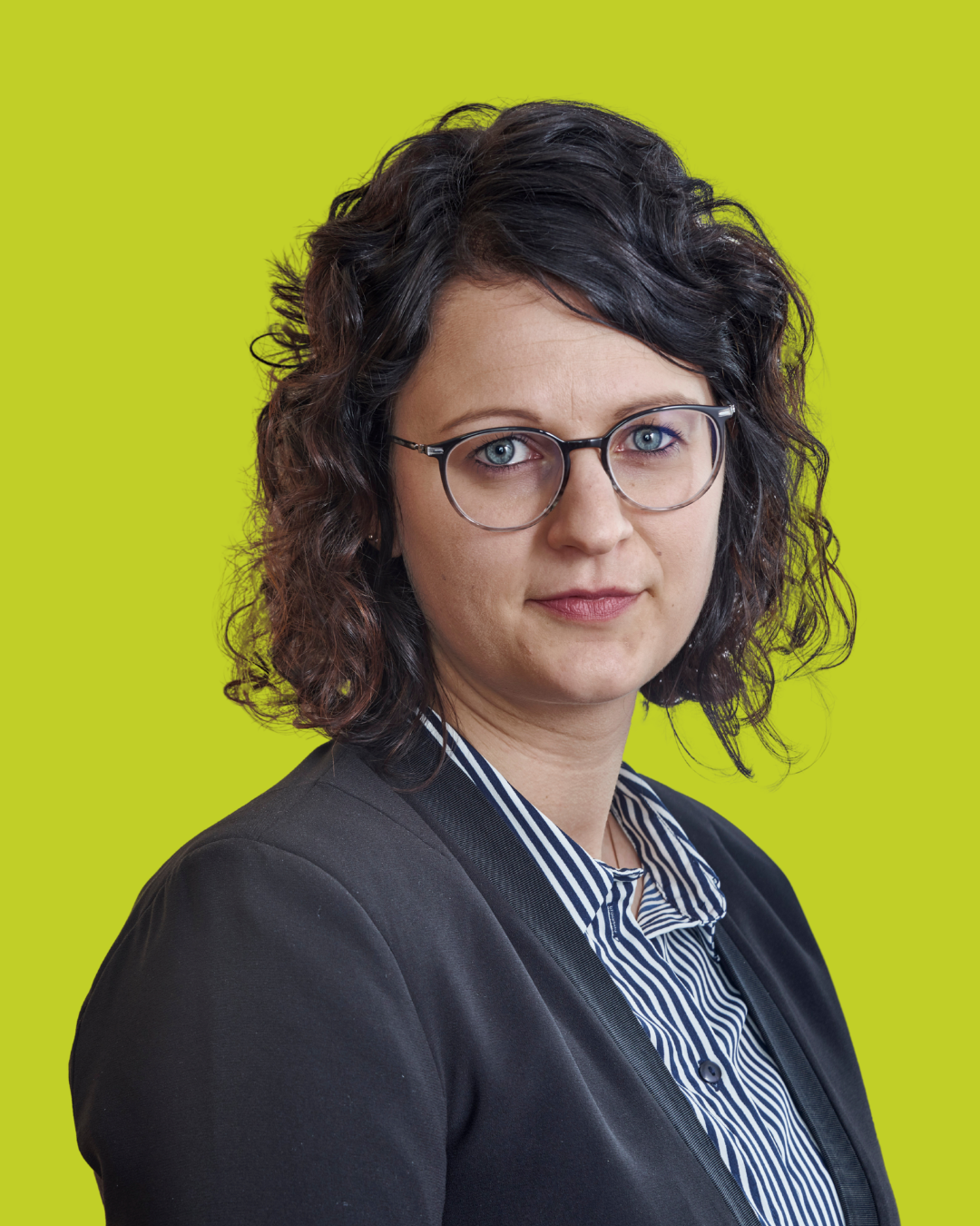 Porträt einer Frau mit lockigen Haaren und Brille im Business-Outfit vor grünem Hintergrund.