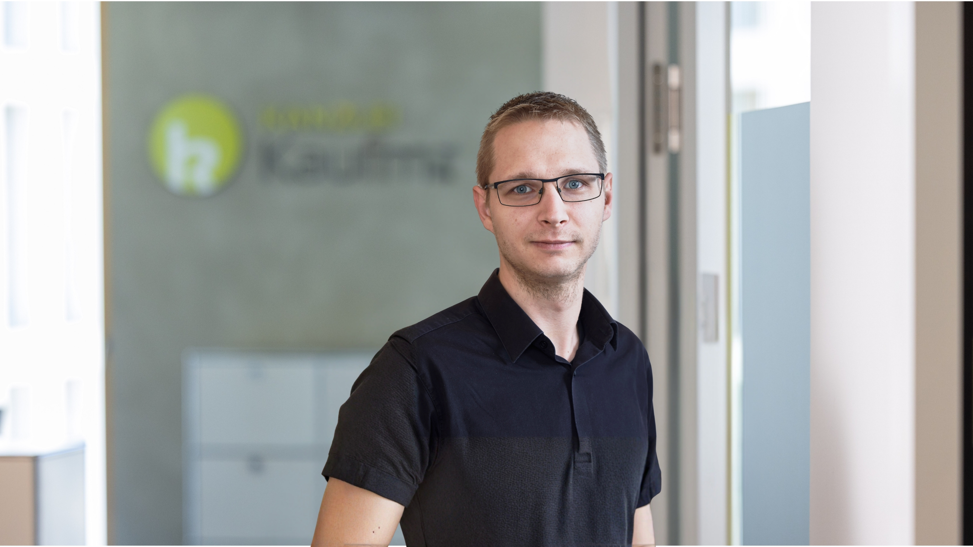 Porträt eines Mannes im schwarzen Poloshirt im Büro vor Glaswand mit Logo.