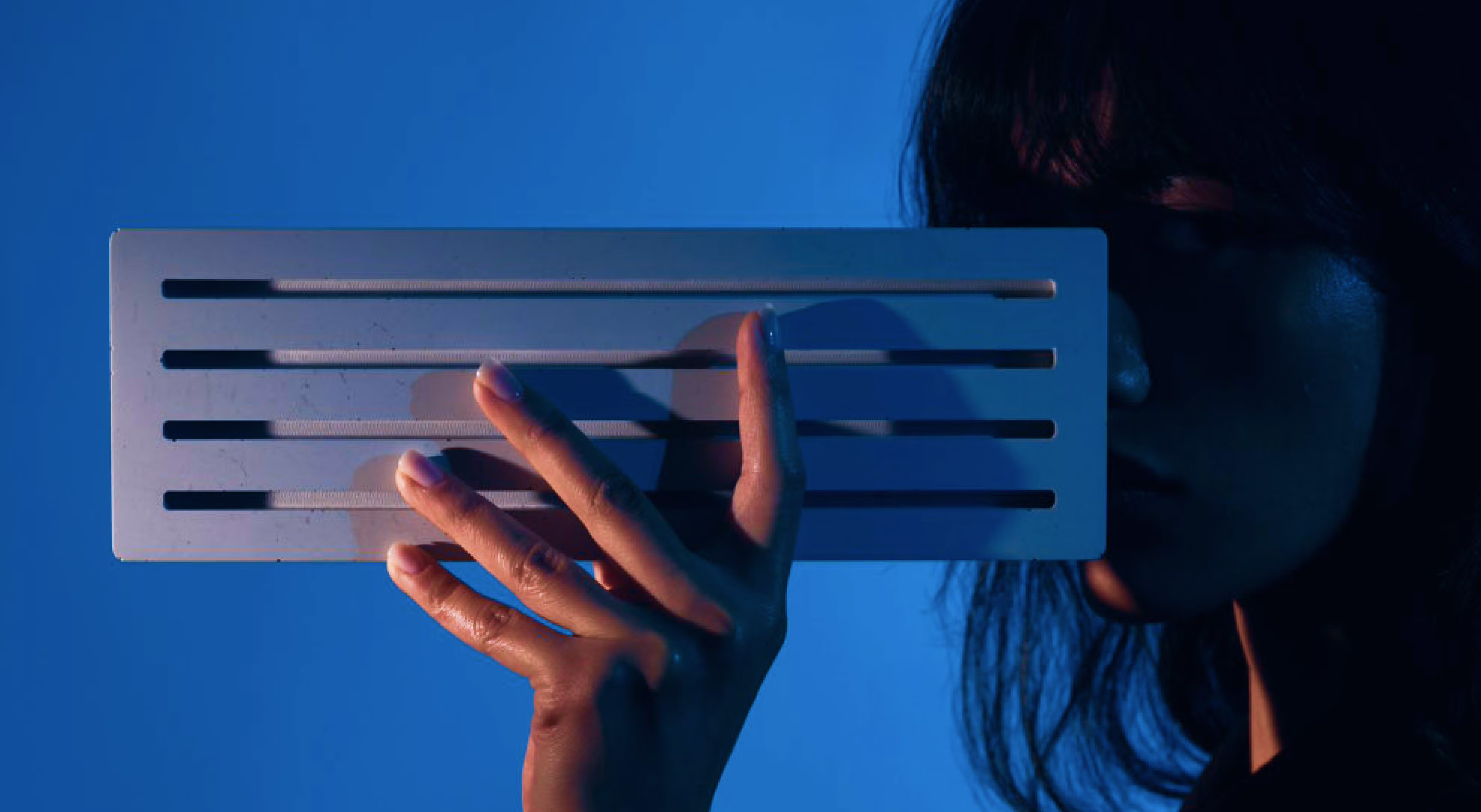 Person holding a white rectangular object with horizontal slots against a blue background.