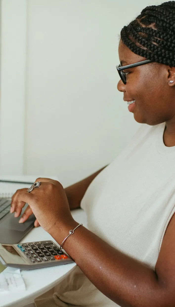 Person with braided hair and glasses using a calculator and laptop at a table.