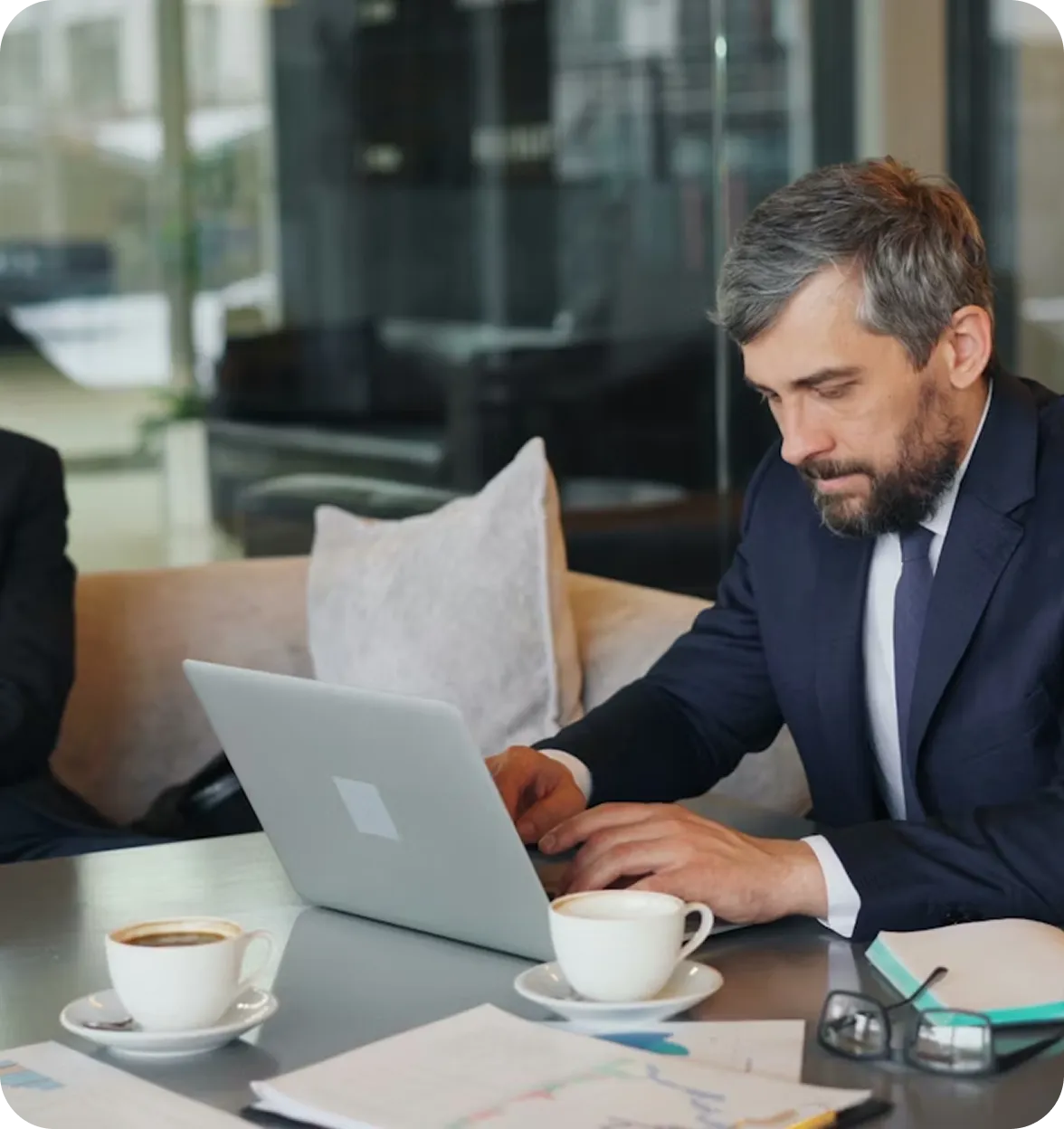 Homme d'affaires concentré travaillant sur un ordinateur portable dans un café avec deux tasses de café, des documents et des lunettes sur la table.