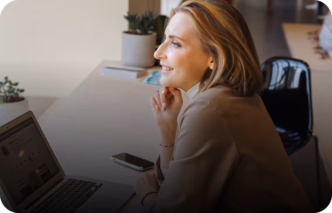 Femme souriante regardant un ordinateur portable ouvert sur un bureau avec un smartphone et des plantes en pot à l'arrière-plan.