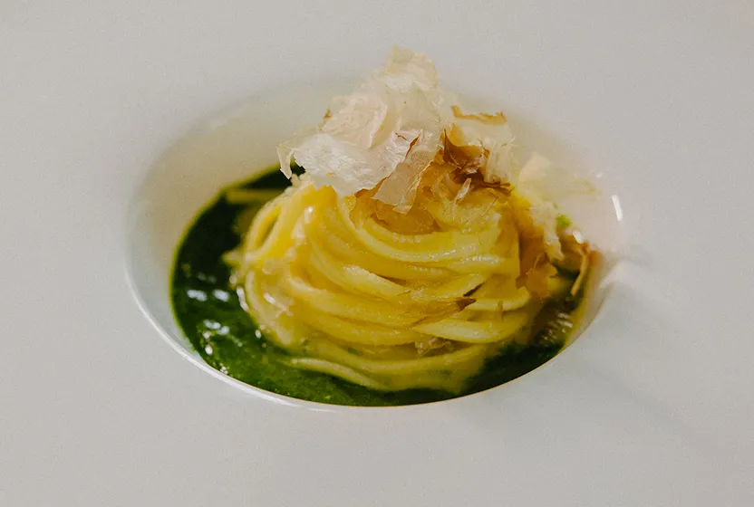 Plate of spaghetti arranged in a nest with green pesto sauce and topped with thin bonito flakes.
