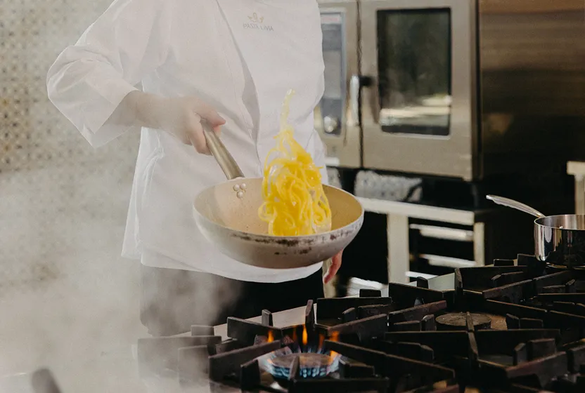 Chef in white Pasta Livia coat tossing pasta in a frying pan over a gas stove flame in a professional kitchen.