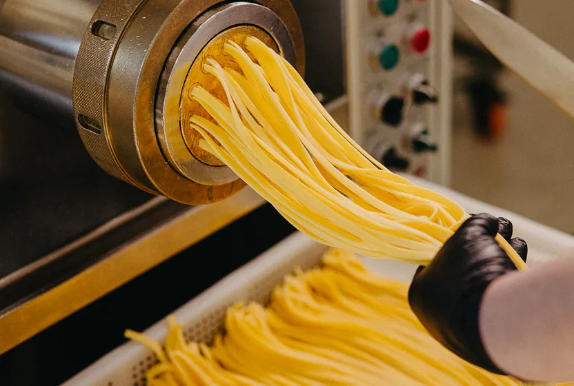 Fresh pasta strands being extruded from a pasta machine, held by a gloved hand.