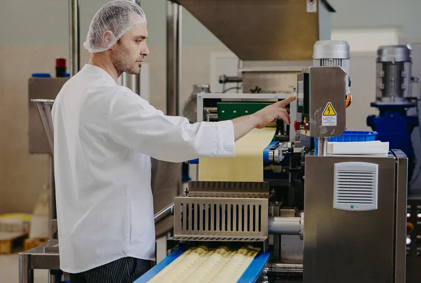 Man in white Pasta Livia coat and hairnet operating a pasta-making machine with sheets of pasta dough on a conveyor belt.