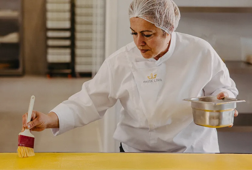 Chef in white Pasta Livia coat and hairnet brushing yellow pasta dough on a table.