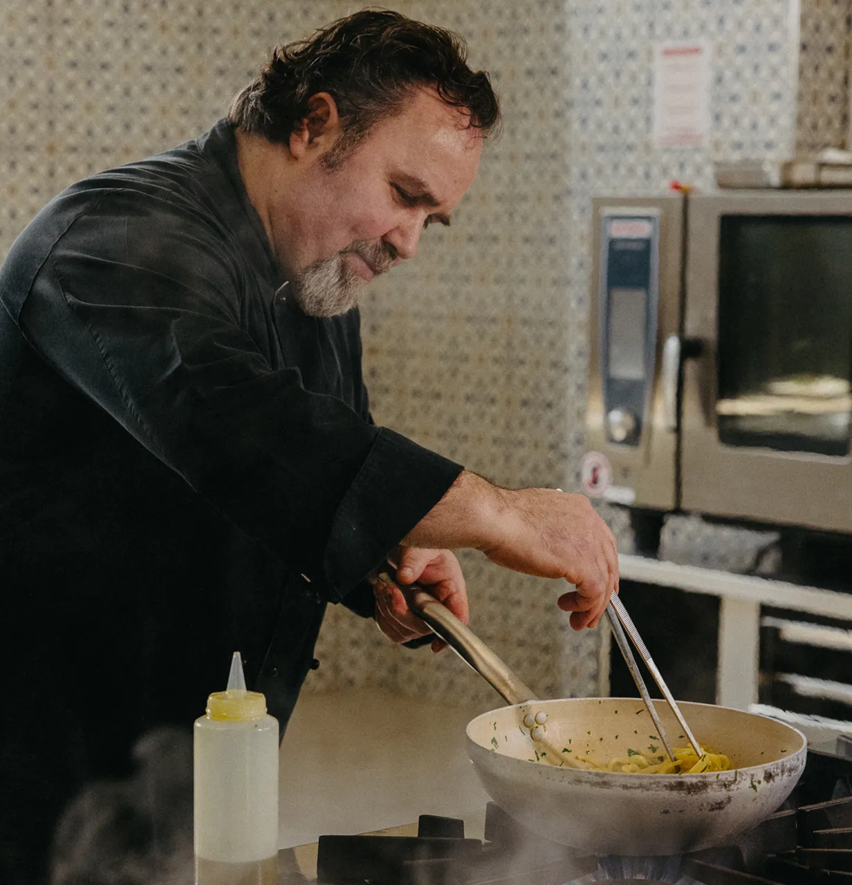 Chef in black Pasta Livia coat cooking pasta in a pan on a gas stove.