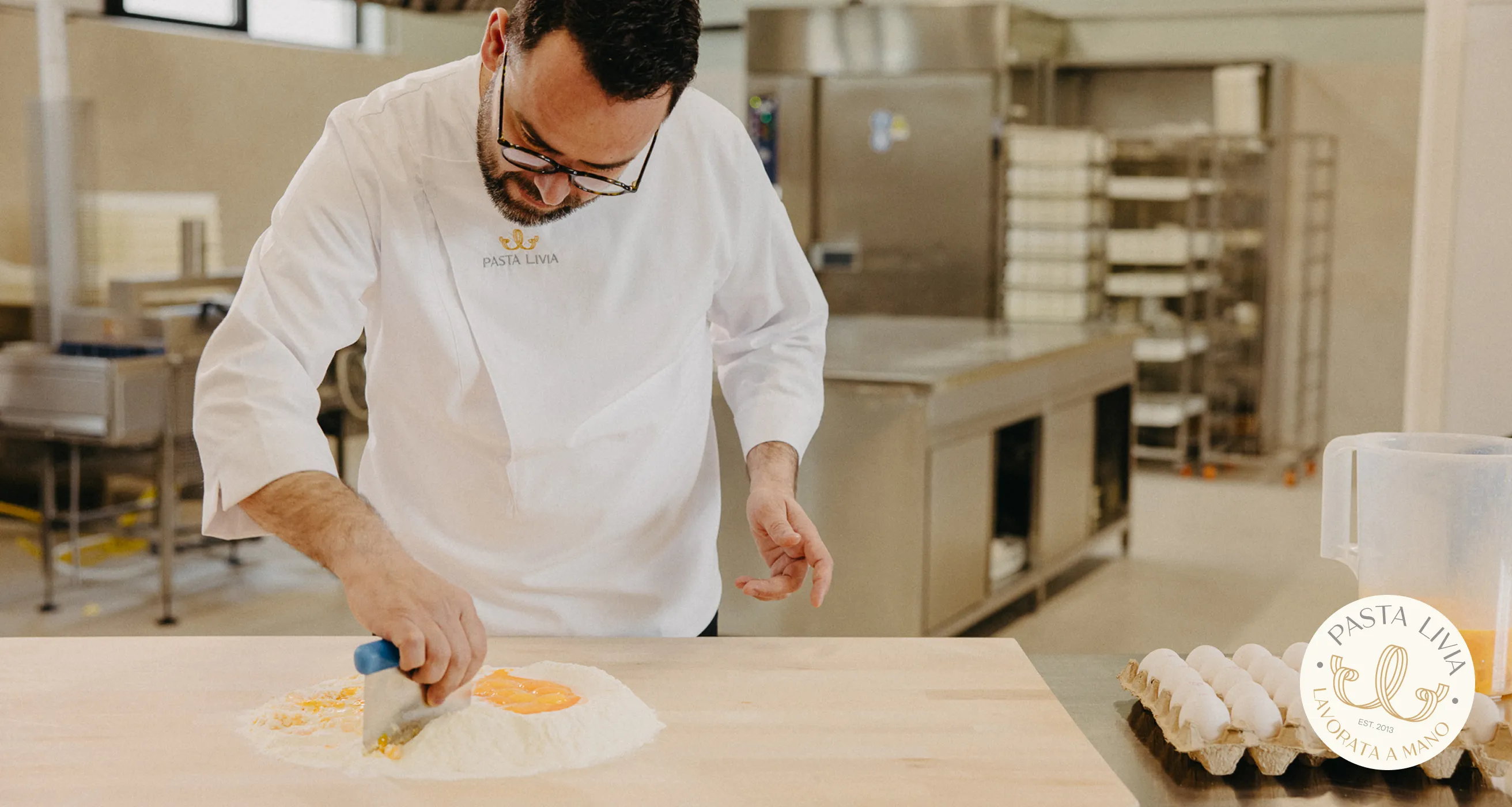 Chef wearing glasses and a white Pasta Livia coat mixing egg yolks with flour on a wooden table in a professional kitchen.