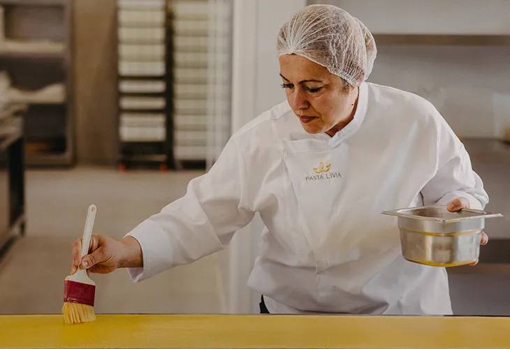 Woman in white Pasta Livia uniform and hairnet handcrafting pasta in a clean kitchen environment.