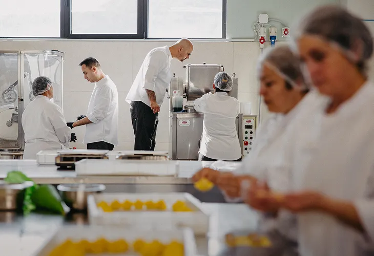 Workers wearing white Pasta Livia uniforms and hairnets preparing food in a commercial kitchen with industrial machines and trays of yellow pasta.