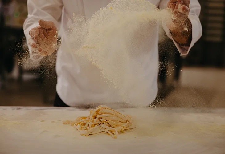 Hands sprinkling flour over fresh pasta dough on a floured surface.