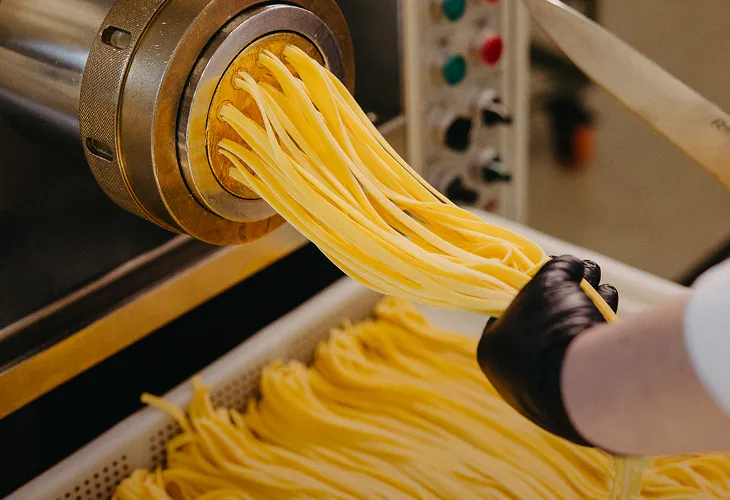 Fresh pasta strands being extruded from a pasta machine, held by a gloved hand.