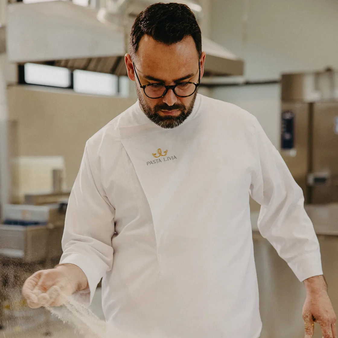 Chef in a white Pasta Livia uniform sprinkling flour in a kitchen.