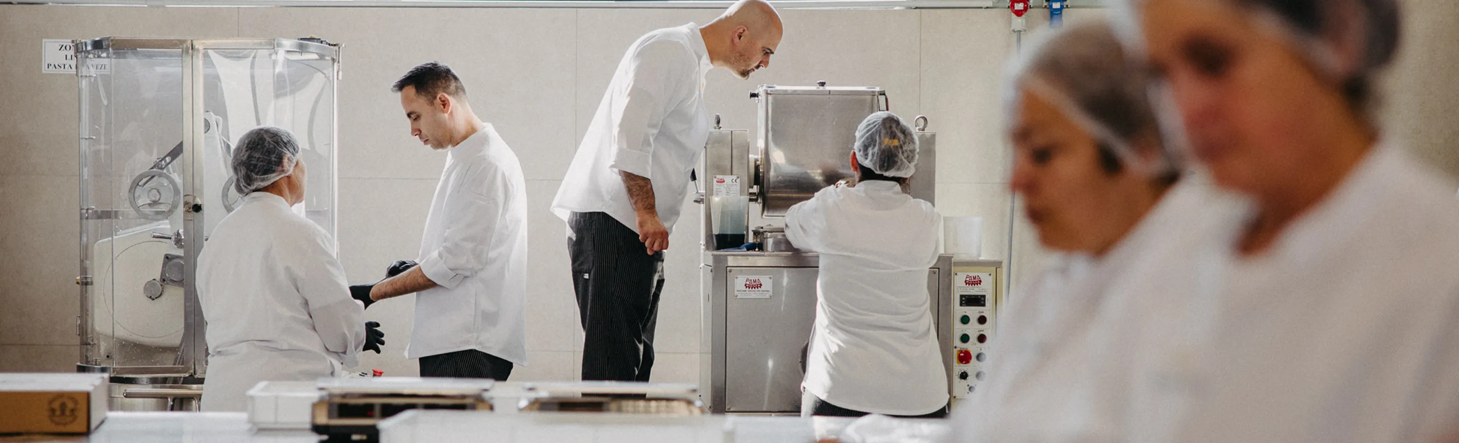People dressed in white Pasta Livia uniforms and hairnets operating pasta-making machinery in a food production facility.