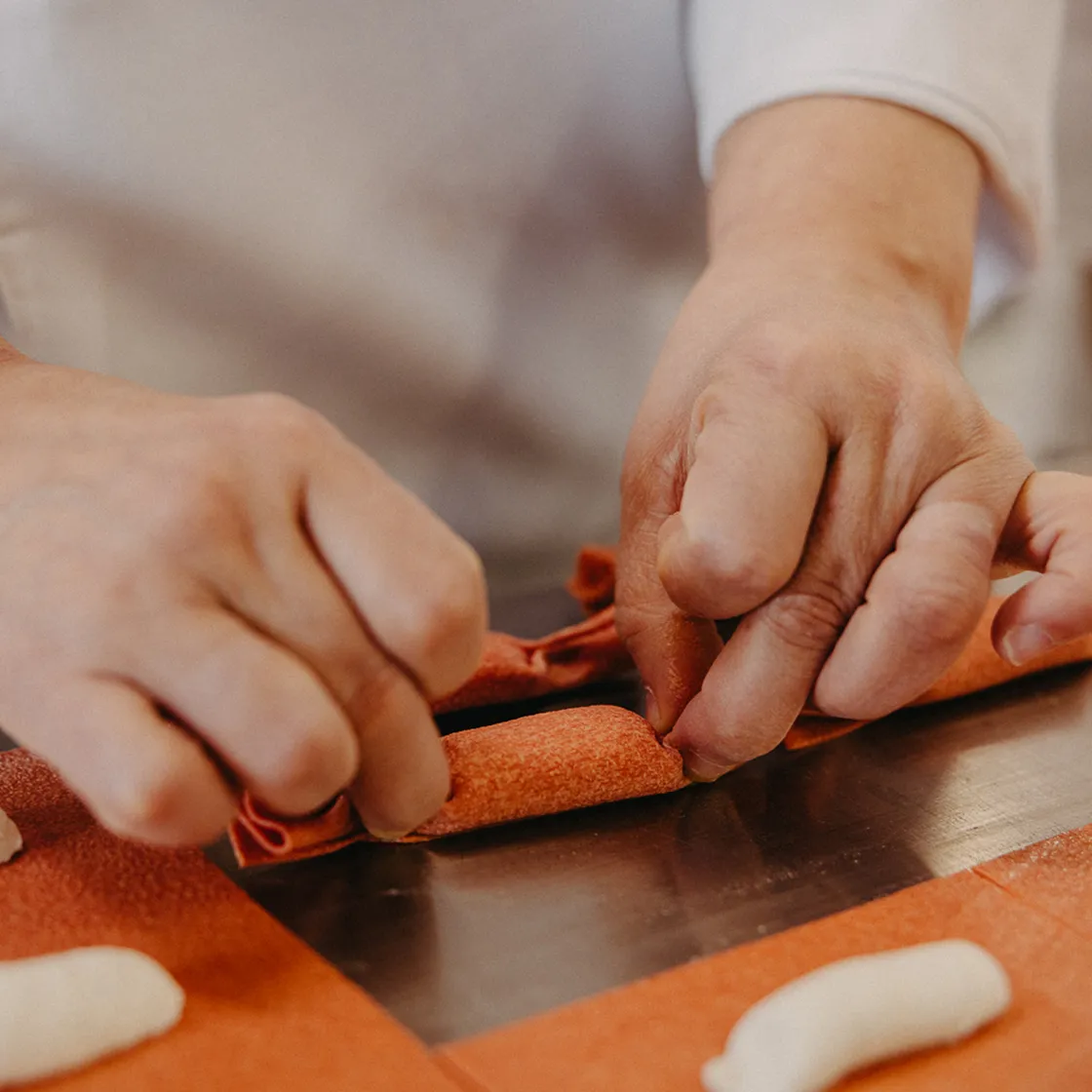 Hands folding red pasta dough over a metal surface, preparing pasta by hand.