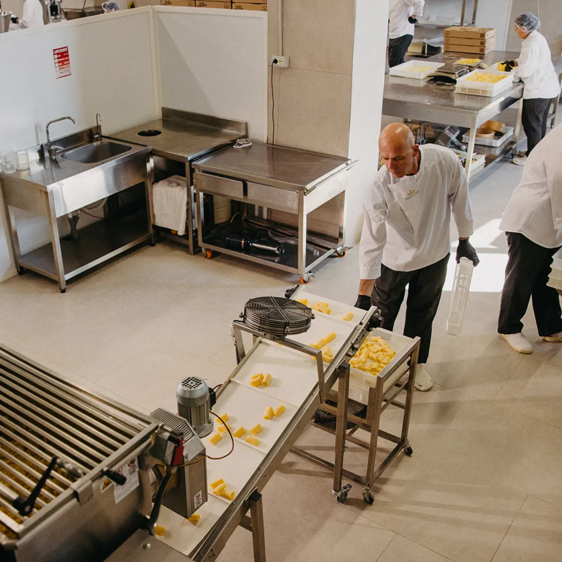 Factory workers in white Pasta Livia uniforms handling pieces of pasta on a conveyor belt in a clean industrial kitchen.
