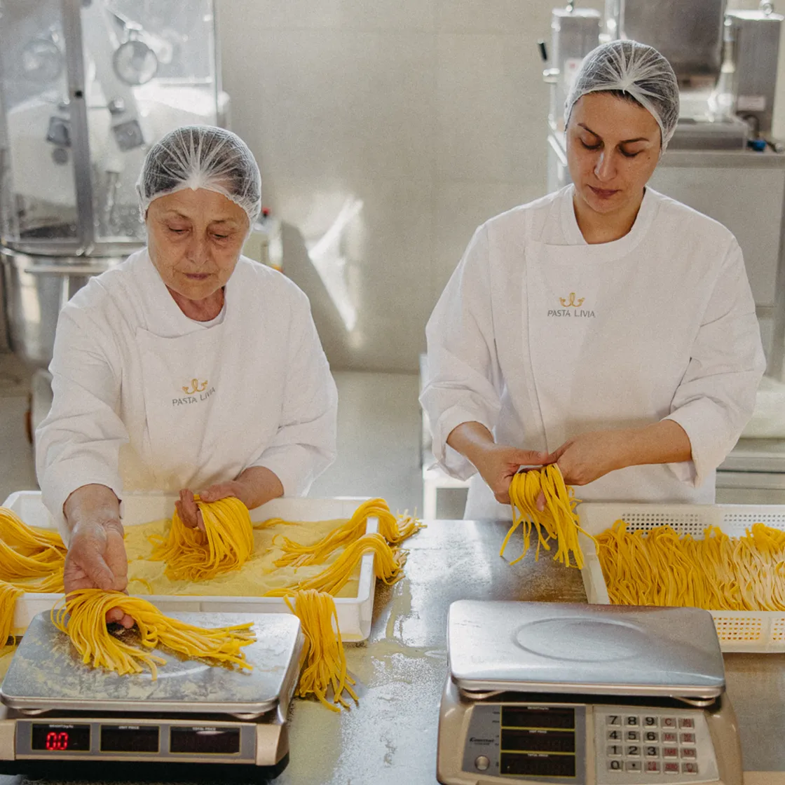 Two women wearing hairnets and white Pasta Livia uniforms weighing and handling fresh yellow pasta strands in a commercial kitchen.