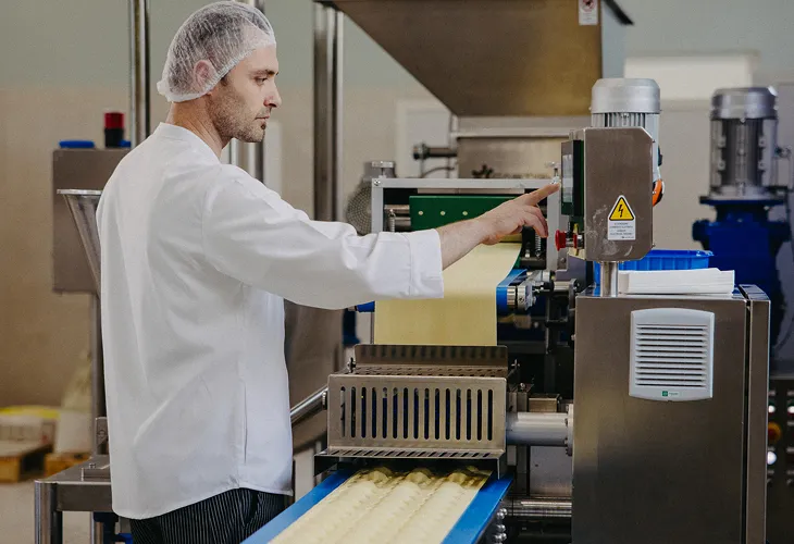 Man in white Pasta Livia coat and hairnet operating a pasta-making machine with sheets of pasta dough on a conveyor belt.
