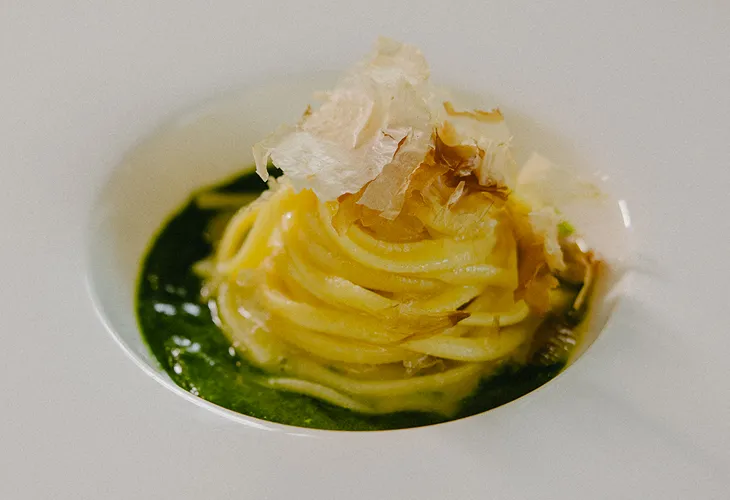 Plate of spaghetti arranged in a nest with green pesto sauce and topped with thin bonito flakes.