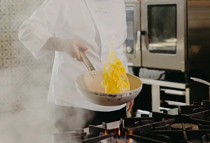 Chef in white Pasta Livia coat tossing pasta in a frying pan over a gas stove flame in a professional kitchen.