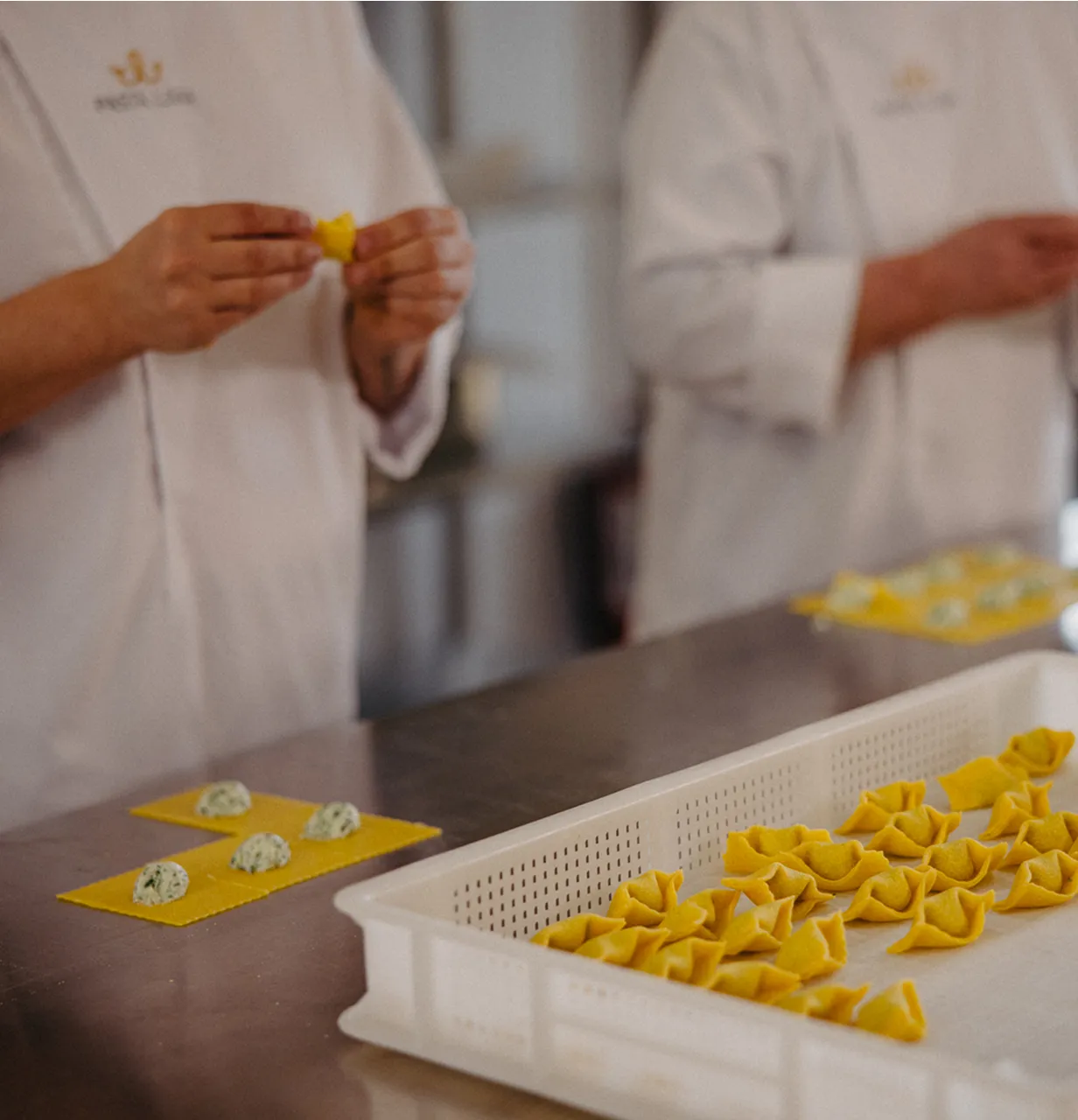 Two chefs in white Pasta Livia uniforms making stuffed pasta on a metal table with a white tray holding completed pasta.