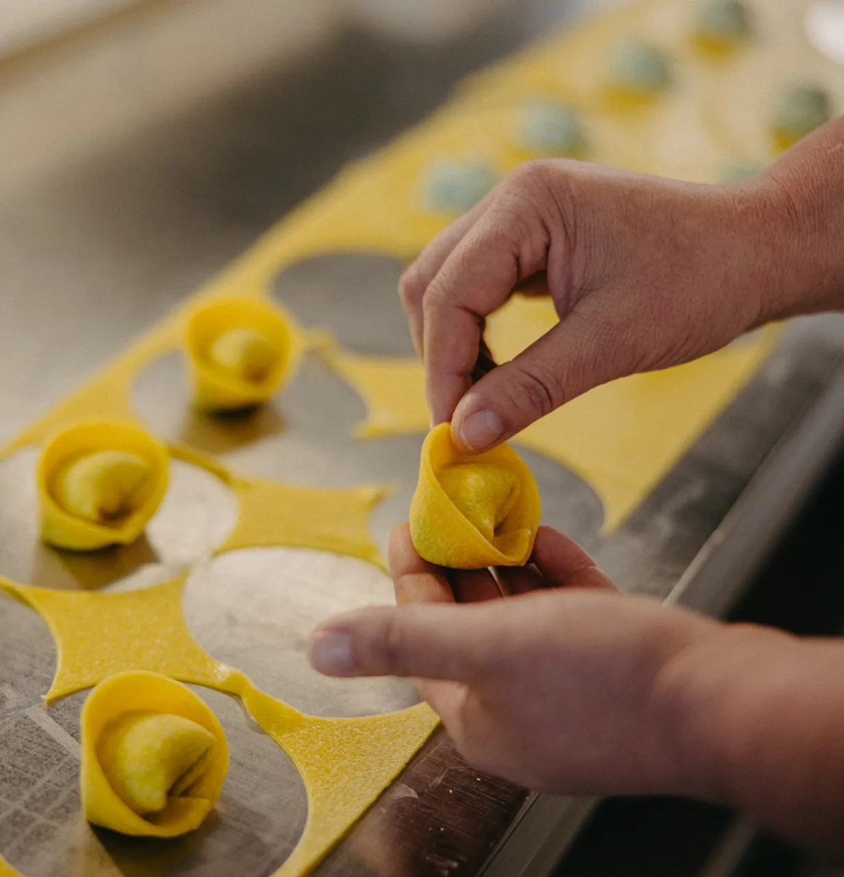 Hands folding fresh yellow tortellini pasta with a filling on a metal surface.