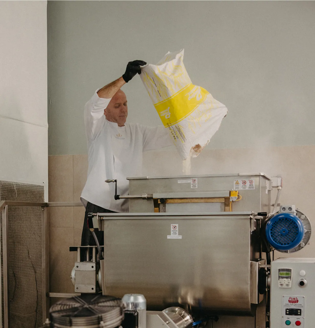 Man wearing a white Pasta Livia uniform and black glove pouring flour from a large bag into an industrial mixing machine.