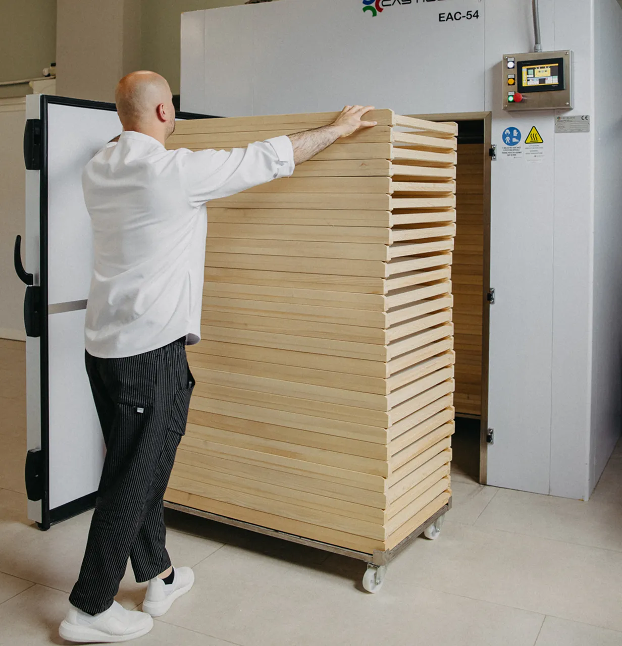 Person in a white Pasta Livia uniform and striped pants pushing a large cart stacked with wooden trays near an industrial oven.