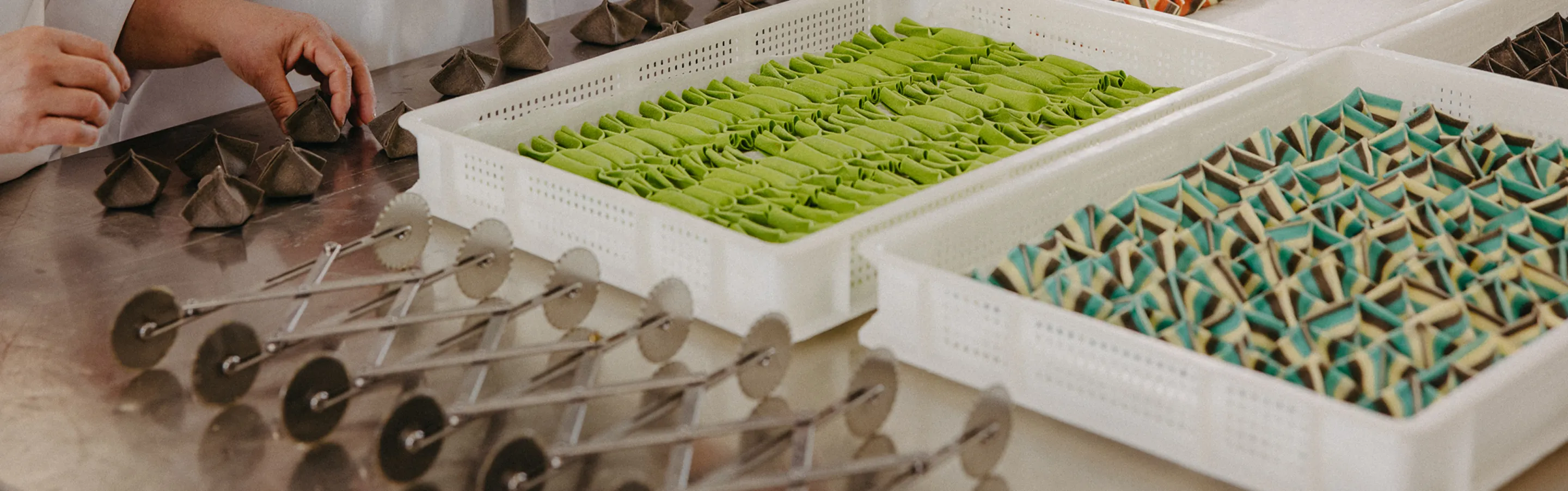 Hands folding and arranging green and multicolored triangular pasta pieces in white trays on a table with several pasta cutters nearby.