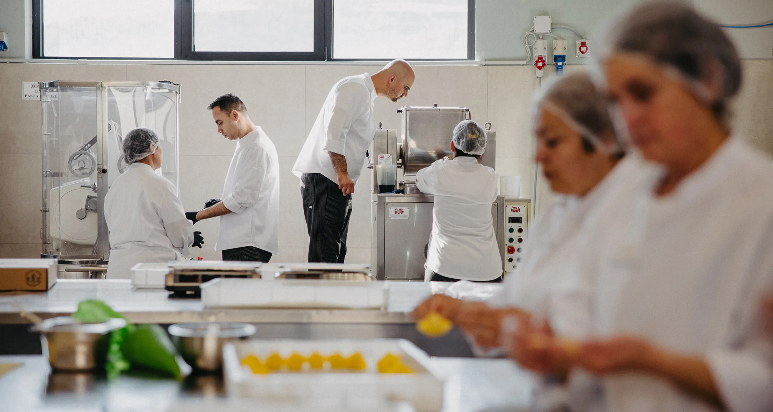 Chefs in white uniforms and hairnets working with pasta-making machines and preparing pasta in a professional kitchen.