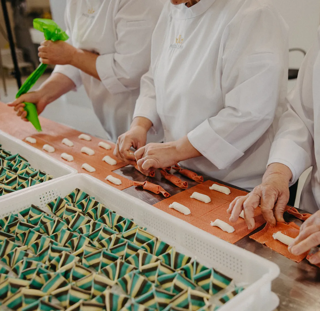 Three people in white Pasta Livia  uniforms preparing and folding red pasta dough with white filling into shapes on a countertop, with trays of striped green and beige folded pasta nearby.