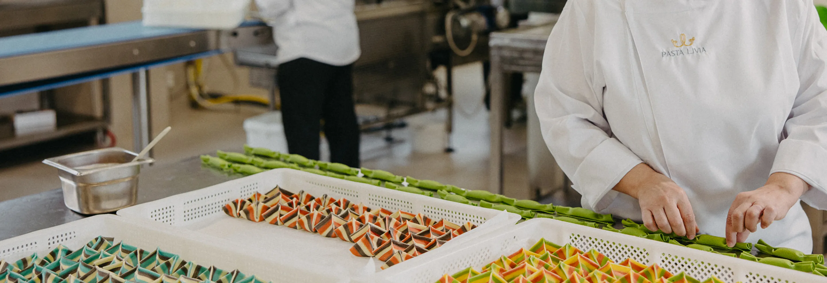Chef in white Pasta Livia uniform arranging colorful handcrafted pasta in trays inside a commercial kitchen.