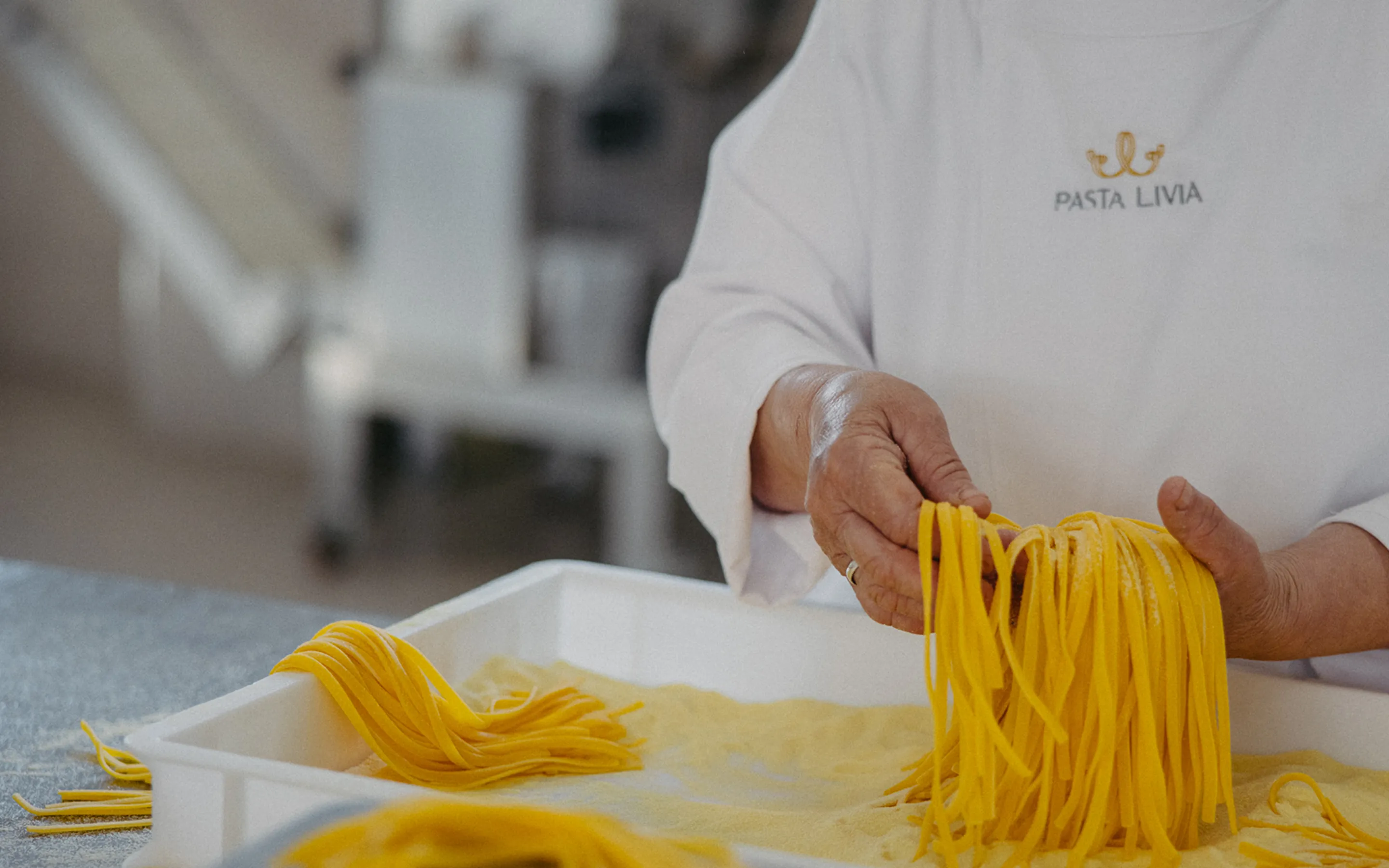 Person wearing a Pasta Livia uniform holding fresh handmade pasta over a tray with more pasta.