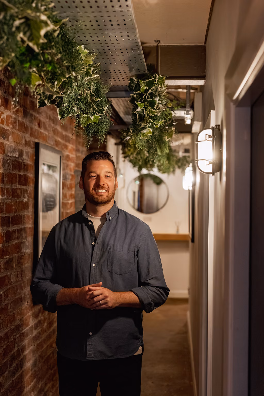 Andy Kelham from Storyhouse Creatives standing in a hallway with exposed brick wall and hanging green plants.
