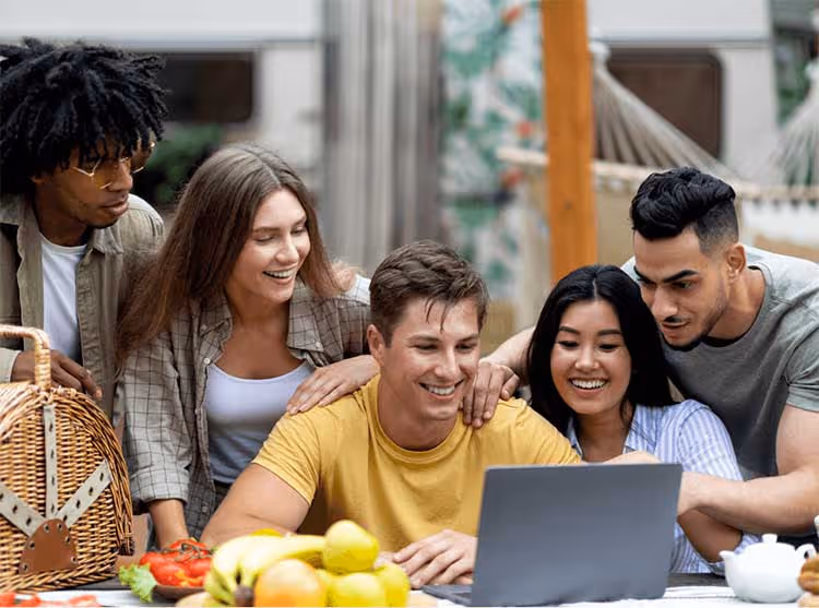 A diverse group of smiling young adults gather around a laptop outdoors, pointing and engaging with content on the screen