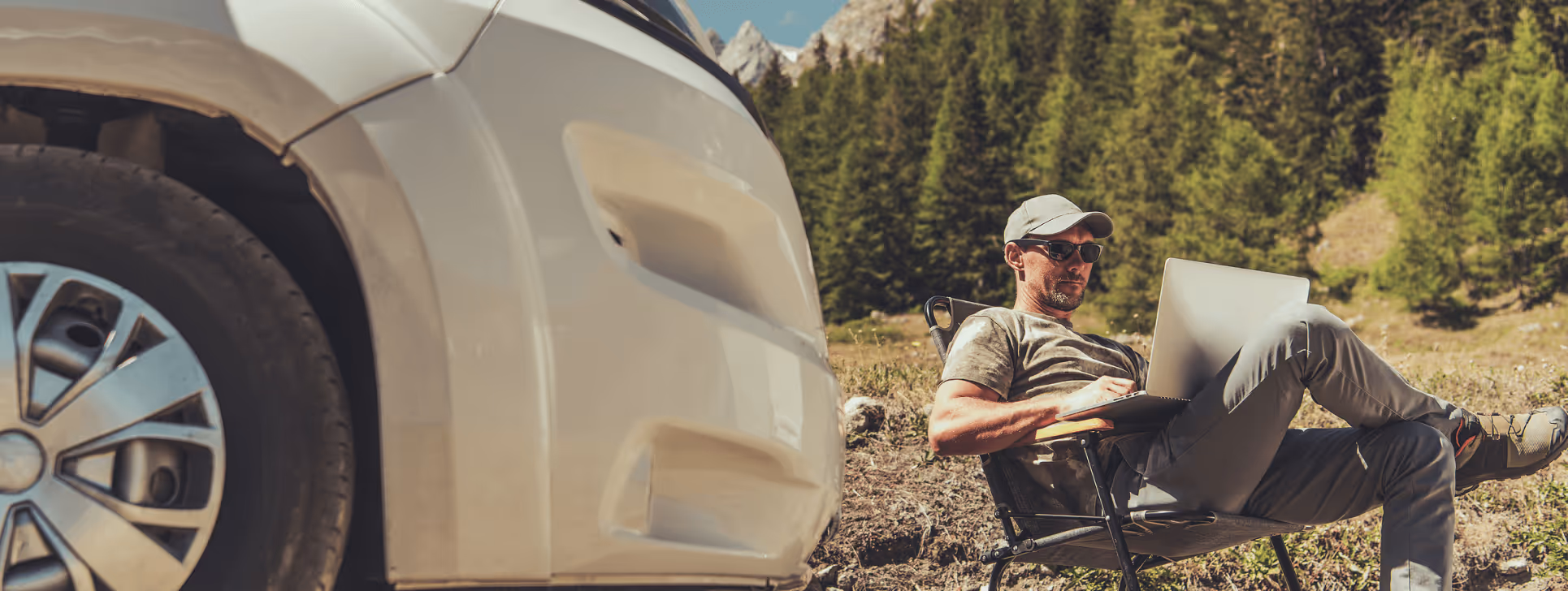 A man in sunglasses and a cap works on a laptop in a folding chair next to a white camper van, surrounded by a mountain and forest landscape, embodying remote work and outdoor adventure.