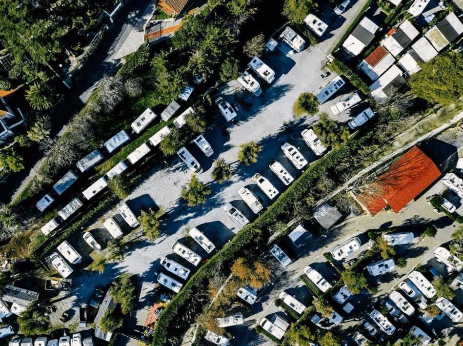 An aerial shot shows numerous white recreational vehicles and trailers parked closely together in an organized layout, surrounded by green trees and some small buildings, suggesting a busy campground or residential RV park.