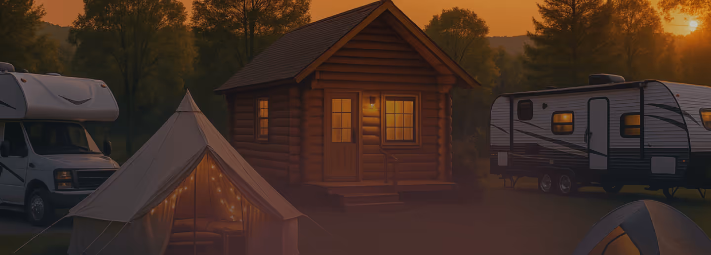 A panoramic view of a cozy campground at sunset, featuring a small wooden cabin with illuminated windows, several RVs, and a decorated tent, surrounded by trees under an orange sky.