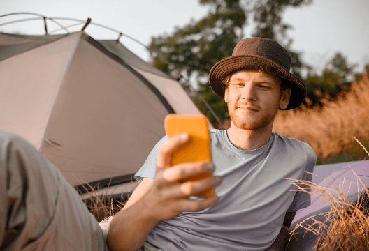 Camper reclining outside a tent while checking a smartphone, illustrating Mass SMS communication to guests.
