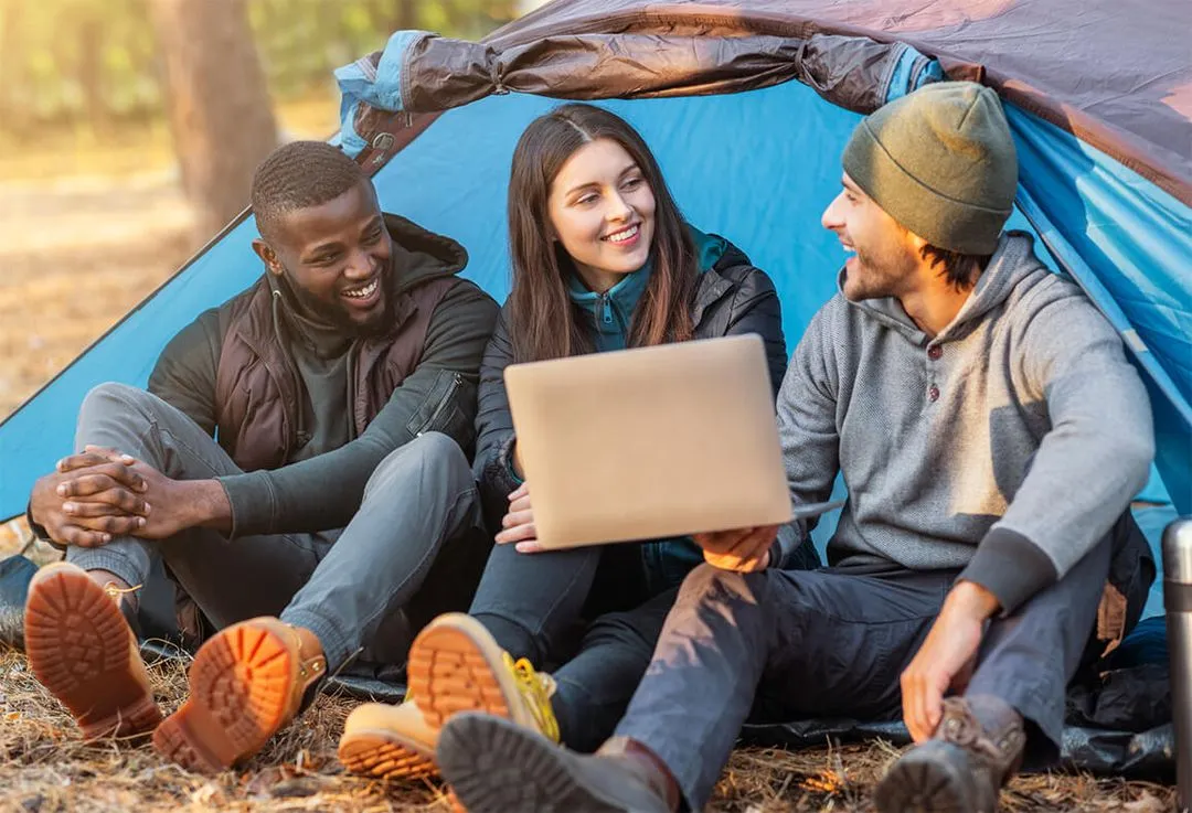 Three friends sitting inside a tent, smiling and looking at a laptop.