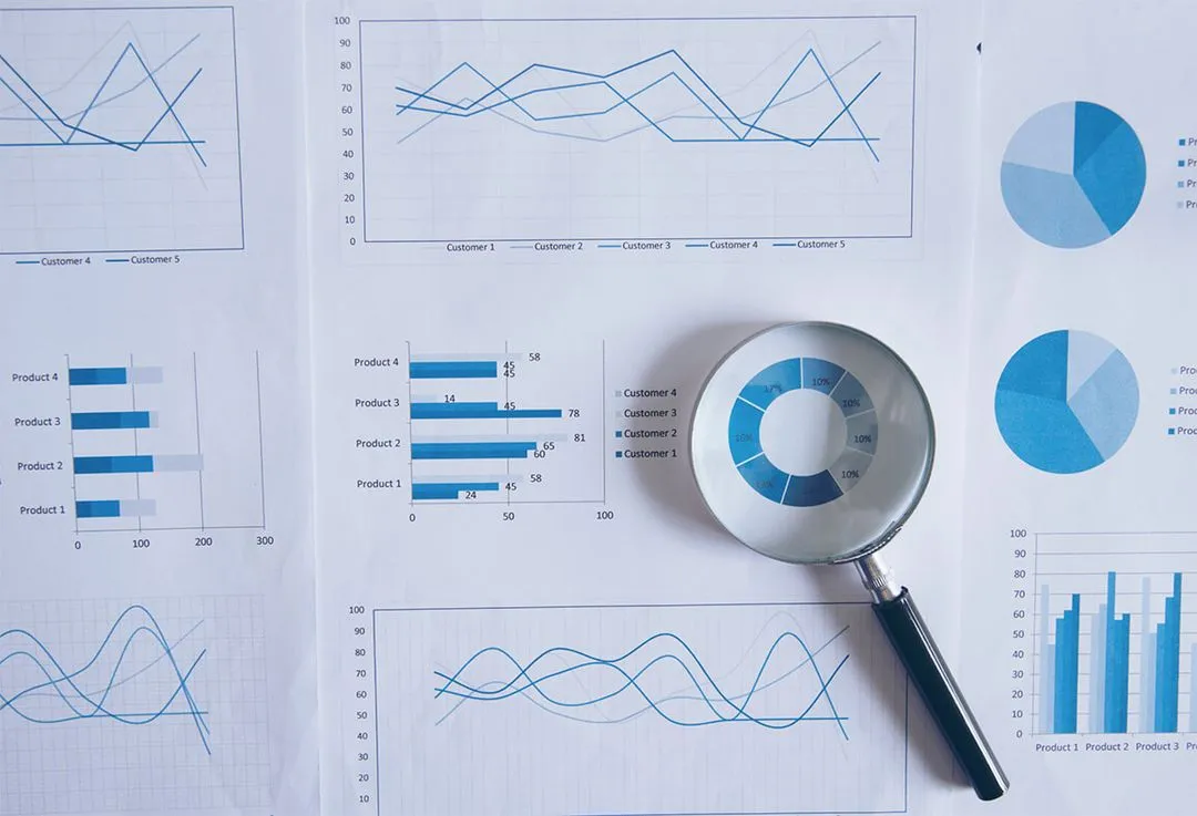Paper printout of line graphs, bar charts and pie charts with a magnifying glass over a donut chart.