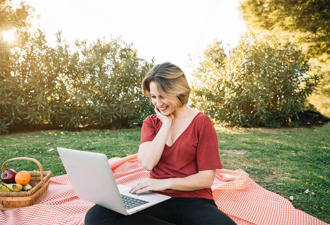 Woman seated on a picnic blanket in a park, smiling at her laptop with a basket of fruit beside her.