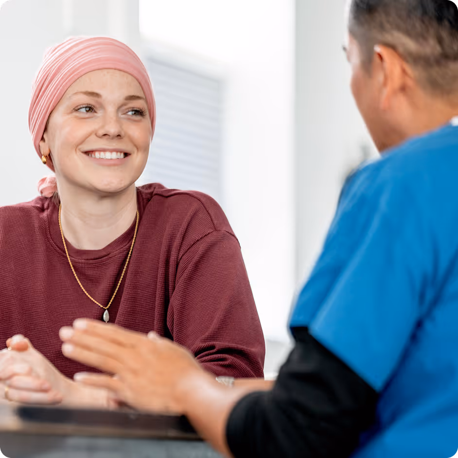 Smiling woman wearing a pink headscarf and maroon sweater talking with a person in blue medical scrubs.
