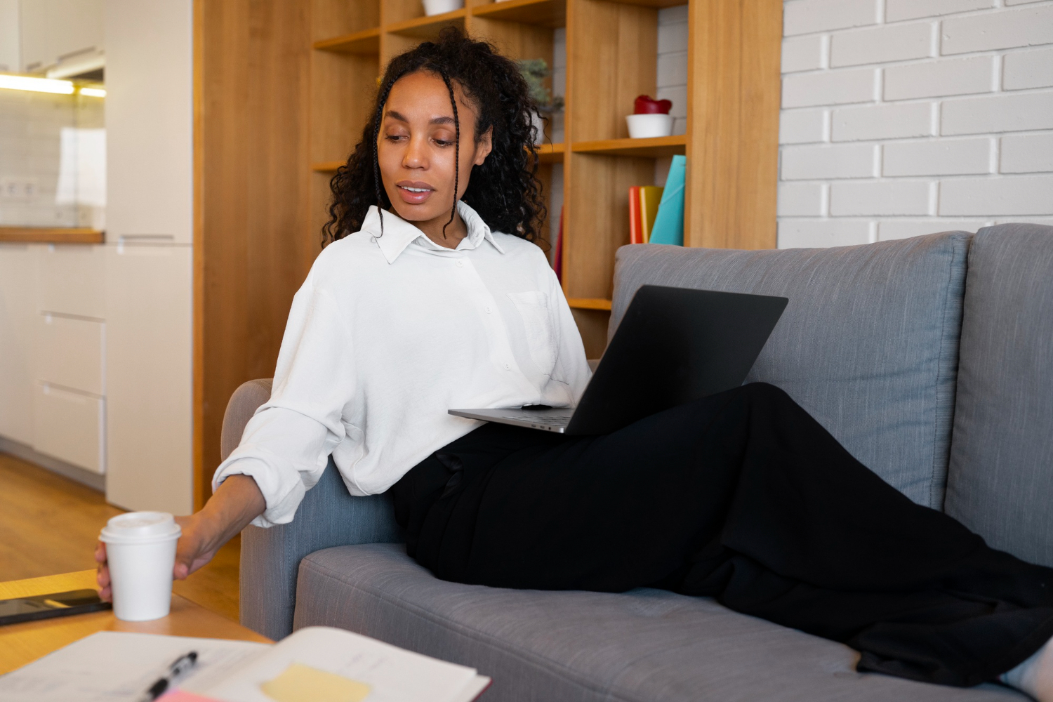 Woman sitting on a couch with a laptop and reaching for a coffee cup, working from home