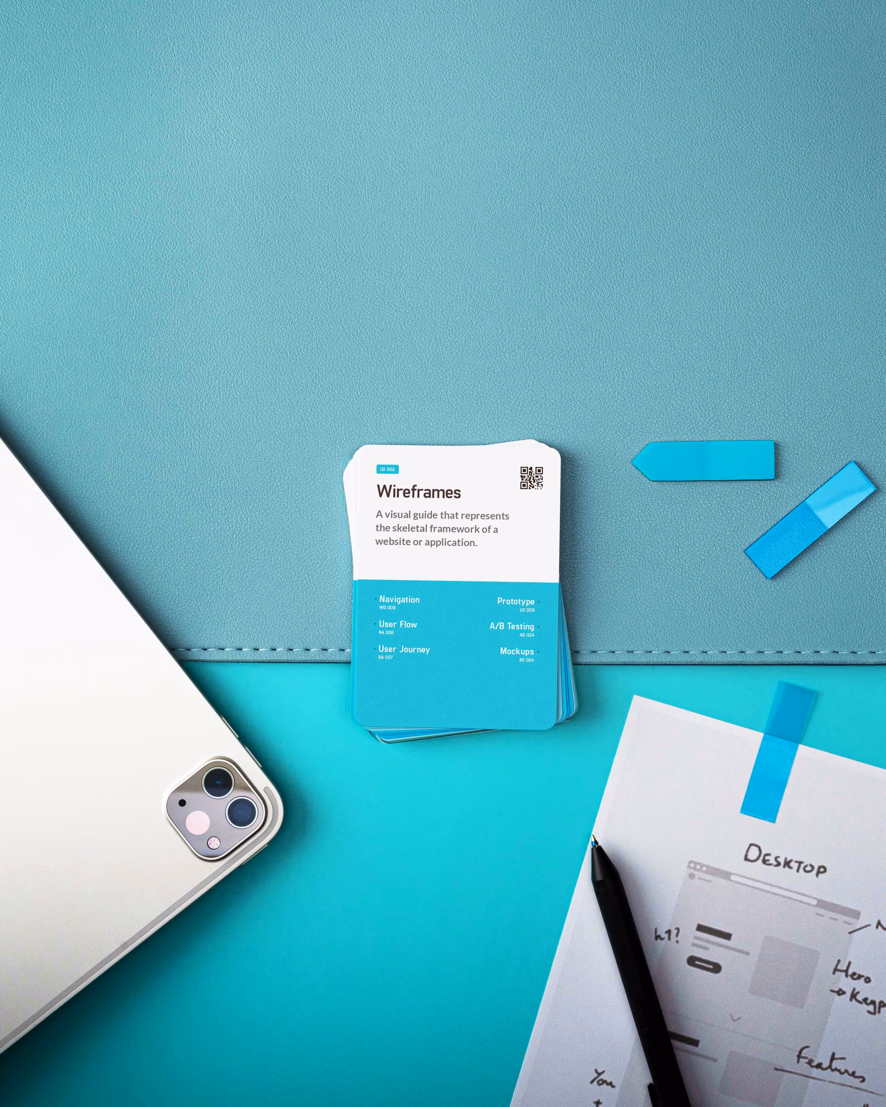 Top view of a workspace with a laptop corner, wireframe cards, sticky notes, handwritten wireframe sketches, and a pen on a teal background.