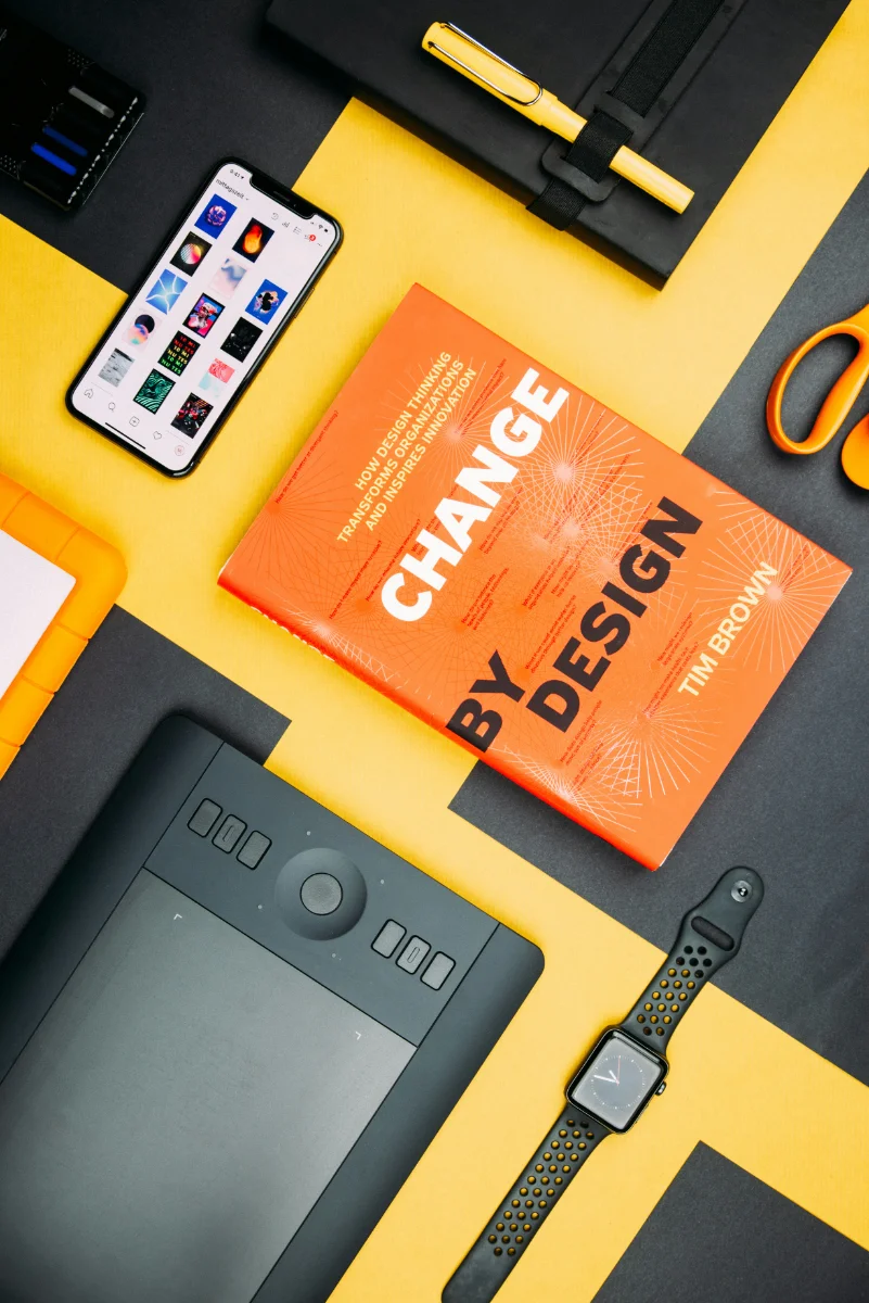 Flat lay of a workspace with an orange book titled 'Change by Design' by Tim Brown, a smartphone showing Instagram, a black digital drawing tablet, a black smartwatch with an orange-accented band, orange scissors, a yellow pen on a black notebook, and a yellow external hard drive on a black and yellow background.